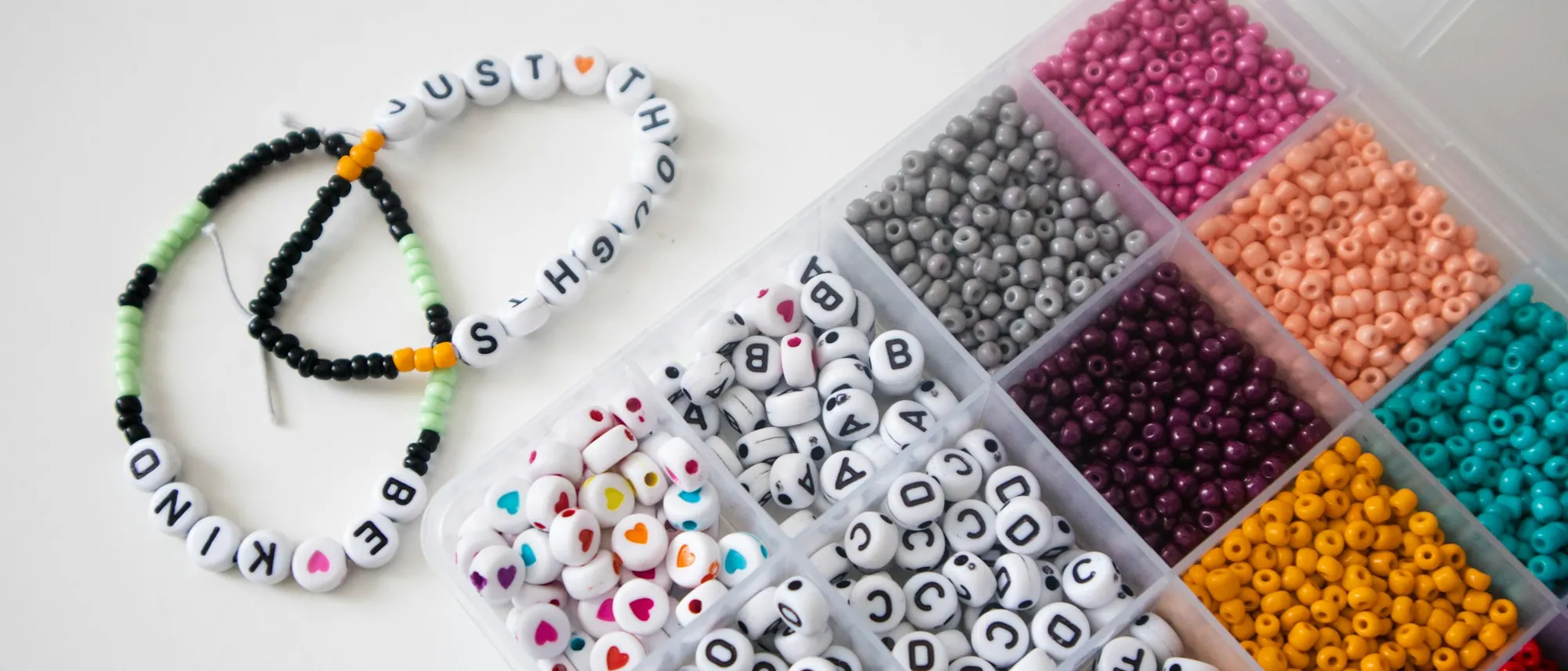 Handmade word bracelets reading BE KIND and JUST THOUGHTS beside a tray of rainbow seed beads and heart-shaped letter beads