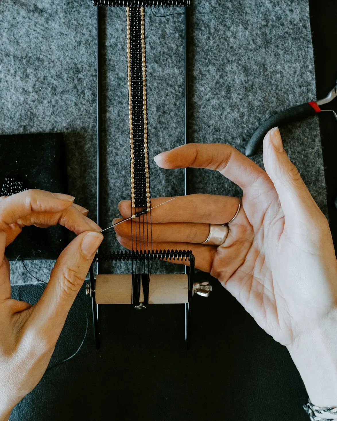 Hands weaving a beaded bracelet on a small loom at a workbench